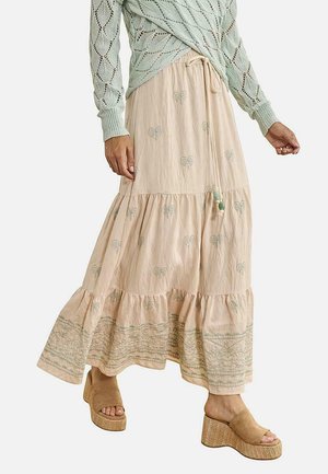 Mujer con falda maxi beige con estampados verdes de palmeras y un suéter de punto verde menta, combinado con sandalias de cuña en tono marrón claro.