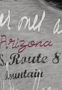 Camiseta de tela gris con texto en varias fuentes, colores y estilos: blanco, negro y burdeos oscuro con un aspecto texturizado y desgastado.