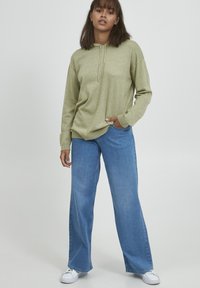 Sudadera de manga larga verde claro con bolsillo frontal y cordón. Combinada con jeans azul de pierna ancha y zapatillas blancas. Textura suave.