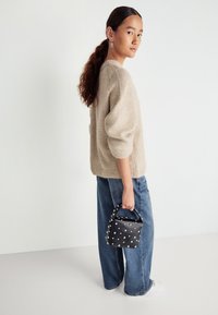 Béžový pletený sveter s vypchávkami na rukávoch, skombinovaný s voľnými modrými džínsami. Osoba drží čiernu kabelku ozdobenú bielymi nitmi.