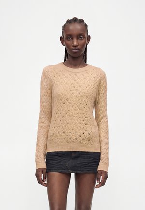 Femme aux cheveux tressés portant un pull en maille beige texturé et une jupe courte en denim foncé, debout devant un fond blanc uni.