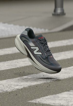 Donkergrijze New Balance hardloopschoen met witte zool in de lucht boven een zebrapad op een geasfalteerde straat.