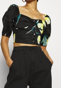 Donna che indossa un top corto a maniche puff nere con stampa di limoni gialli e pantaloni neri a vita alta, mani nelle tasche, collana d'oro e orecchini a cerchio.