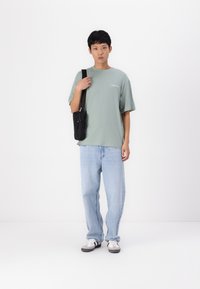 Tricou din bumbac verde deschis cu logo, croială lejeră. Blugi din denim albastru deschis, croială liberă. Geantă de umăr neagră, adidași gri și albaștri.