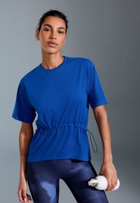 Tricou sportiv albastru cu mânecă scurtă și talie strânsă, asortat cu colanți negri cu un model de nori, ținut cu o sticlă de apă albă.