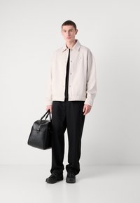 Giacca bomber beige chiaro, maglietta nera, pantaloni neri, sneakers nere, con una borsa nera strutturata con manico superiore e dettagli in zip.