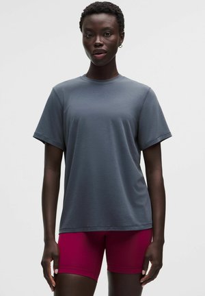 Persona joven con el pelo rizado y corto, vistiendo una camiseta gris holgada y pantalones cortos rojos ajustados, de pie frente a un fondo claro y liso.