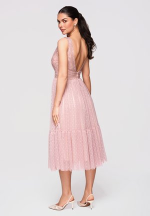 Kobieta o ciemnych włosach związanych nisko w kucyk, ubrana w różową, bezrękawnikową sukienkę midi z koronki i tiulu, z głębokim dekoltem w kształcie litery V z tyłu oraz białe sandały na obcasie z paskiem z tyłu.