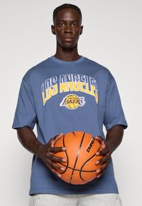 Siva kratka majica s kratkimi rokavi z grafiko "LOS ANGELES LAKERS" v modri in rumeni barvi. Model drži oranžno košarkarsko žogo.