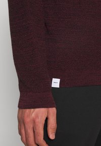 Bordo pletený svetr s žebrovanými manžetami a texturovaným vzorem. Na dolní straně je viditelná bílá etiketa s nápisem „MAKIA".