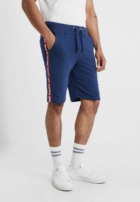 Shorts en coton bleu marine avec taille à cordon, poches latérales et bande rouge avec texte. Associés à des baskets blanches et des chaussettes rayées.
