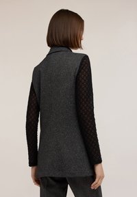 Gilet grigio con una finitura testurizzata, maniche sheer nere a contrasto con un motivo circolare, e un design di colletto classico.