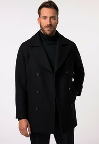 Abrigo de lana negro de doble botonadura con grandes solapas, bolsillos frontales y seis botones; llevado sobre un suéter de cuello alto negro.
