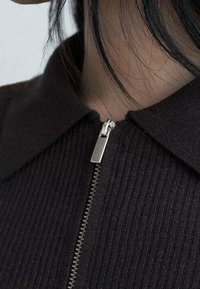 Haut marron foncé à côtes avec un col pointu et un détail de fermeture éclair argentée au niveau du cou, mettant en valeur le tissu texturé et les lignes épurées.