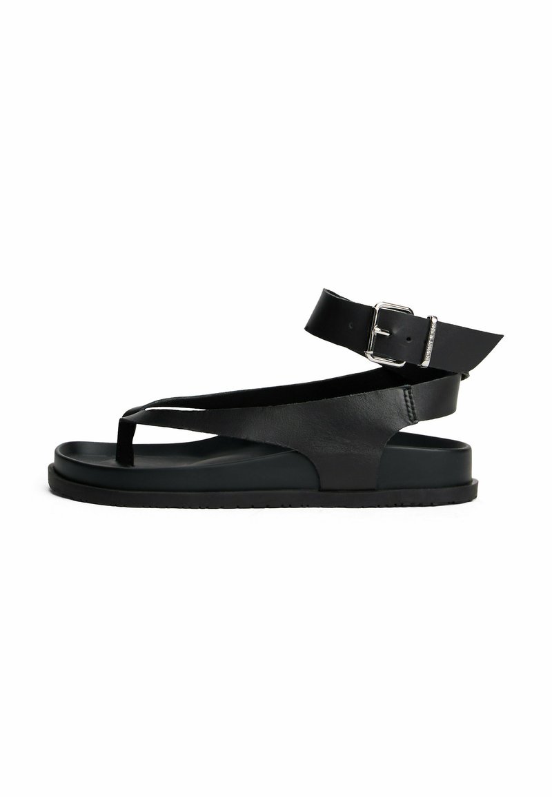 Sandalo in pelle nera con design aperto sulla punta, cinturino alla caviglia con fibbia e suola piatta spessa. Texture liscia e silhouette minimalista.