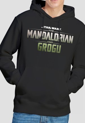 Person trägt schwarzen Hoodie mit dem Schriftzug „Star Wars The Mandalorian and Grogu“ in Grau und Grün, Hände in der Fronttasche, blaue Jeans sichtbar.