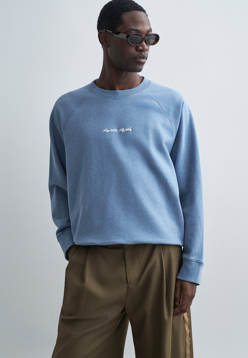 Sudadera de algodón azul claro con mangas raglán, con el logotipo bordado en blanco "Maison Kitsuné" en el pecho, combinada con pantalones marrones.