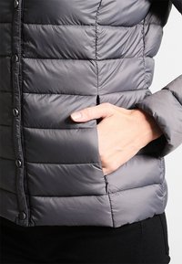 Grå puffet jakke med quiltet design, der har knaplukning og sidelomme. Glat tekstur, letvægt og slank silhuet.