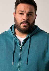 Tyrkysová mikina na zip s kapucí, předním zipem a měkkou texturou. Bílé tričko pod ní. Detailní záběr horní části těla a obličeje muže.