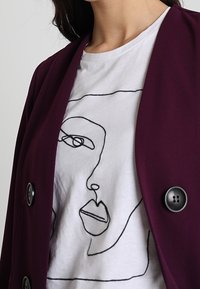 Bordeauxblazer med synliga knappar lager-på-lager över en vit t-shirt med en svart linjetecknad ansikte. Slät tygtextur.