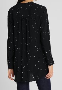 Blouse noire à manches longues avec motif d'étoiles argentées, coupe décontractée, poignets à boutons et un empiècement froncé à l'arrière. Texture de tissu douce.
