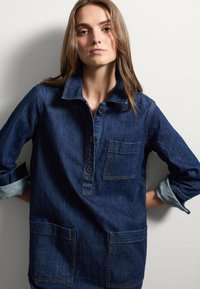 Donkerblauwe denim shirt met een kraag, knoopsluiting en twee zakken aan de voorkant. Heeft opgestroopte mouwen met lichtblauwe accenten.