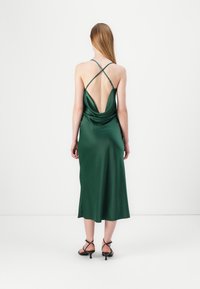 Rochie din satin verde cu spate drapat, având bretele încrucișate. Țesătura are o textură fină și o lungime până la jumătatea gambei, completată cu pantofi cu toc negri.