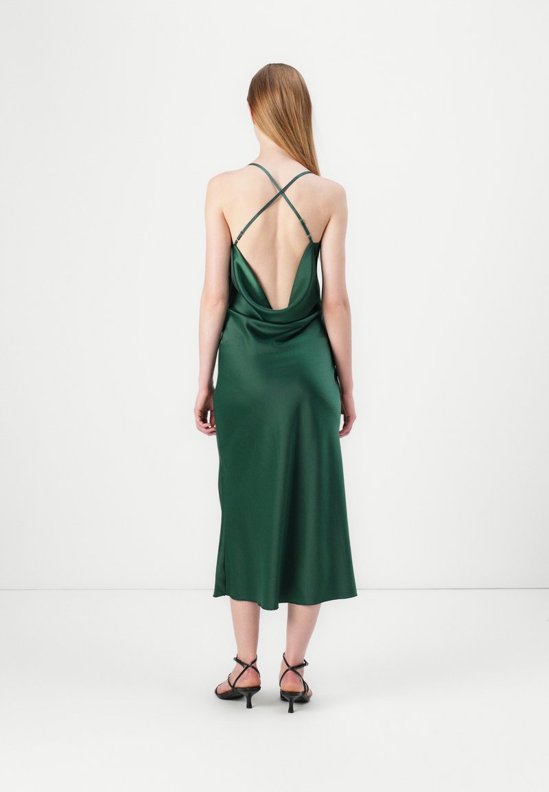 Rochie din satin verde cu spate drapat, având bretele încrucișate. Țesătura are o textură fină și o lungime până la jumătatea gambei, completată cu pantofi cu toc negri.