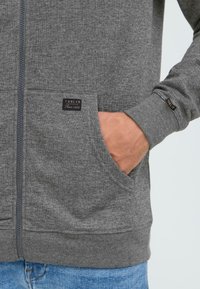 Siva zip-up dukserica napravljena od mekanog materijala, s prednjim džepom i kontrastnim šavovima. Oznaka je vidljiva s imenom brenda i godinom osnivanja.