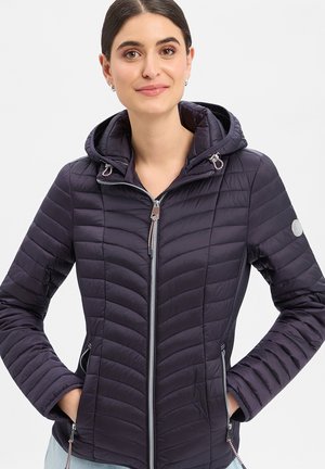 Vrouw met een donkere gewatteerde jas met capuchon, ritssluiting aan de voorkant, ritssluitingen aan de zijzakken en een logopatch op de linker mouw, handen in de zakken.