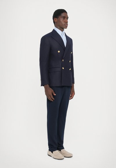 Blazer bleu marine à double boutonnage avec des boutons dorés, associé à une chemise rayée bleu clair et un pantalon sombre, complété par des mocassins beiges.