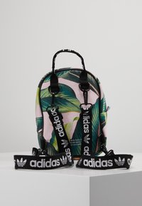 Mały plecak z różowym tłem, zdobiony zielonymi wzorami liści, czarnymi paskami i białym logo "adidas", oraz uchwytem na górze.