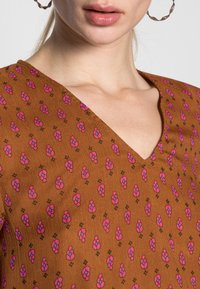 Brauner Stoff mit pinkem Blumenmuster und dunklen Akzenten, mit V-Ausschnitt-Design. Die strukturierte Oberfläche verleiht dem gesamten Look Tiefe.