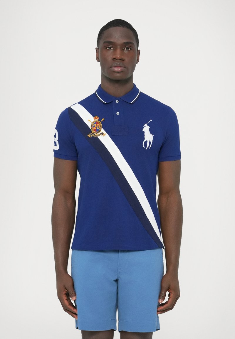 Tengerészkék galléros póló fehér csíkkal és emblémával, rövid ujjú. Világoskék rövidnadrággal viselve. Ujján logó és a 3-as szám látható.