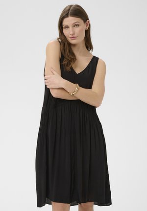 Junge Frau mit langem braunem Haar, die ein ärmelloses schwarzes Kleid und ein goldenes Armband trägt, steht vor weißem Hintergrund mit verschränkten Armen.