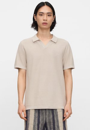 Jonge persoon met halflang zwart haar draagt een beige polo met korte mouwen en gestreepte grijs-beige broek, staat stil.