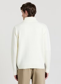 Mann mit kurzen braunen Haaren, der einen weißen Rollkragenpullover und beige Hosen trägt, mit dem Rücken zu einer schlichten hellen Hintergrund.