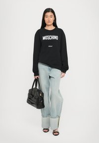 Sudadera negra con texto blanco "MOSCHINO", vaqueros de pierna ancha en azul claro con detalles en el dobladillo, bolso negro y sandalias negras de punta abierta.