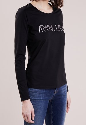 Osoba ubrana w czarną koszulę z długim rękawem z napisem "ARMANI JEANS" i niebieskie dżinsy, stojąca na tle jednolitego jasnego tła.