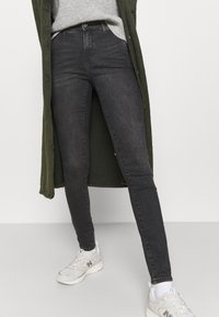 Jeans negros ajustados de mezclilla, con un diseño clásico de cinco bolsillos, combinados con un suéter gris y un cárdigan largo verde.