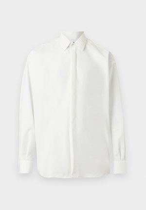 Chemise blanche à manches longues avec col pointu et poignets boutonnés, présentée à plat sur un fond gris clair.