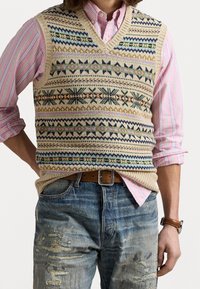 Stickad beige väst med flerfärgade geometriska mönster, bärs över en randig rosa och vit skjorta. Har en v-ringning och ribbad kant.