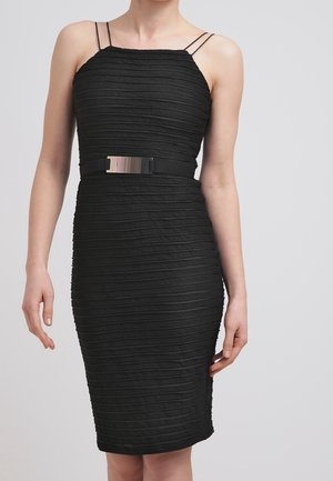 Femme portant une robe noire ajustée texturée avec de fines bretelles doubles et une ceinture noire avec une boucle rectangulaire brillante.