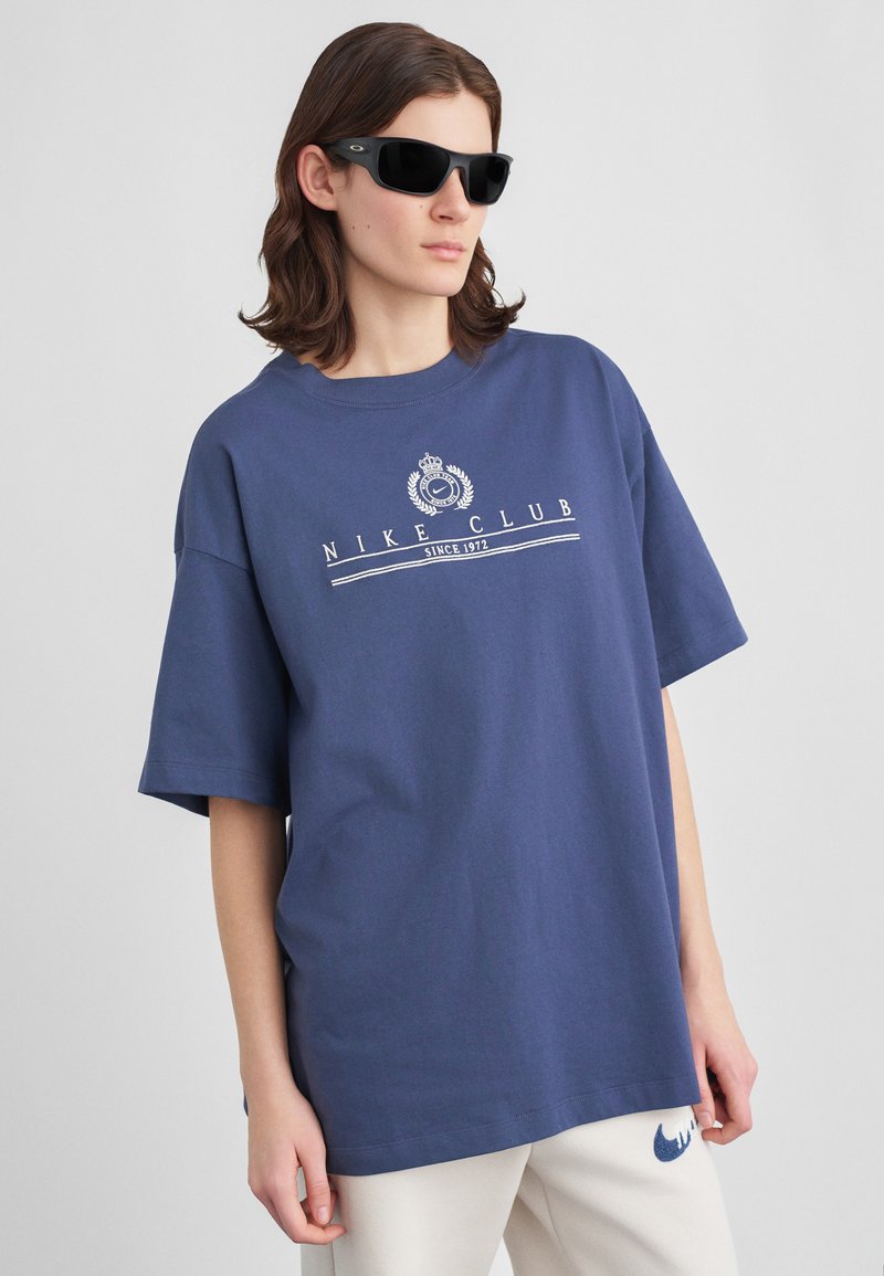 Άτομο που φοράει μια υπερμεγέθη μπλε t-shirt Nike Club, λευκά παντελόνια με μπλε λογότυπο και μαύρα γυαλιά ηλίου, στέκεται μπροστά σε απλό ανοιχτό γκρι φόντο.