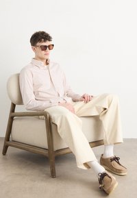 Camicia a righe rosa e bianca con bottoni, pantaloni ampi crema, scarpe in suede marroni, occhiali da sole marroni, seduto su una sedia imbottita beige.