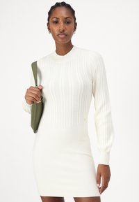 Abito in maglia color crema chiaro, con costine verticali, collo alto e maniche lunghe a sbuffo. L'abito ha una silhouette aderente.