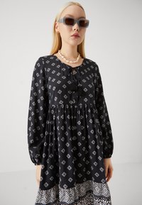 Μαύρο φόρεμα με λευκά διαμαντένια σχέδια, με στενή μέση, φουσκωτά μανίκια και ροτόντα στο τελείωμα. Διακοσμημένο με λαιμόκοψη τύπου κλειδιού και φούντες.