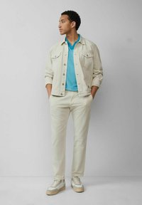 Helles beiges Denim-Jacket, türkisfarbenes Polo-Shirt, cremefarbene Hose und weiße Sneakers mit beigefarbenen Sohlen. Das Modell steht vor einem neutralen Hintergrund.