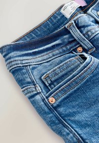 Jeans din denim de un albastru deschis, cu o textură țesută vizibilă, cusături portocalii și accente cu nasturi din cupru. Include o mică buzunar frontal.