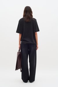 Maglietta nera a maniche corte abbinata a jeans in denim scuro a vestibilità ampia. Una borsa tote marrone è tenuta nella mano destra, vista da dietro.